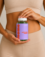 A person in beige activewear holds a bottle of Momo Apple Cider Vinegar dietary supplement. The bottle has a lavender purple label with orange text and a white cap, showing green capsules inside.
