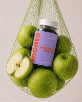 A bottle of momo apple cider vinegar with a purple label sits inside a green mesh bag filled with whole and sliced green apples, photographed against a plain light beige background.