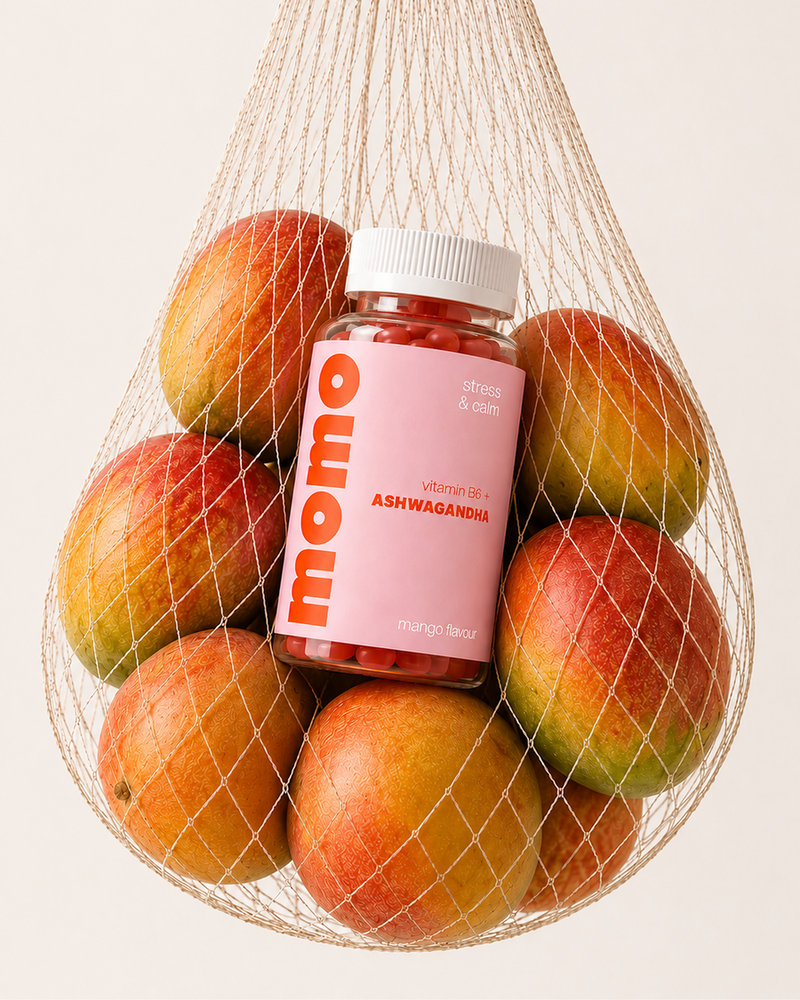 A bottle of Momo Vitamin Ashwagandha in mango flavor, nestled inside a white mesh bag filled with fresh mangoes, on a light beige background.