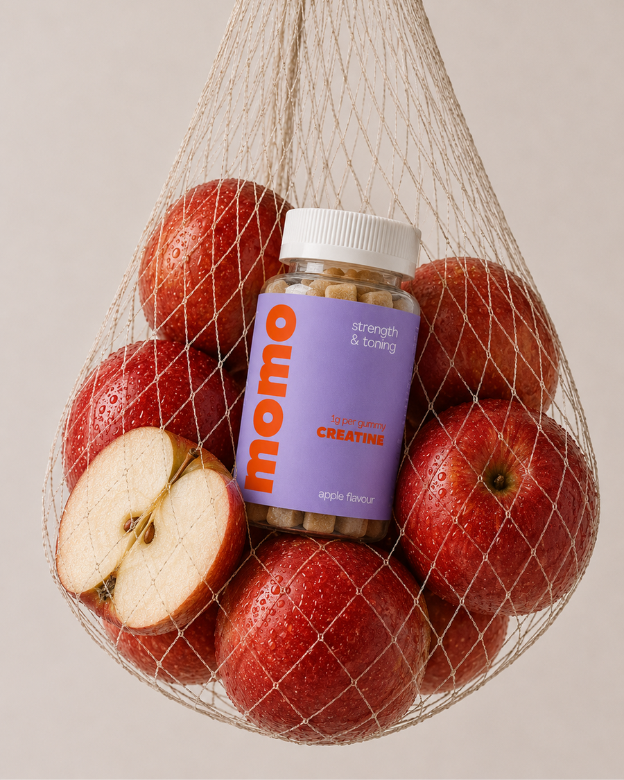 A bottle of momo Strength & Toning Creatine in apple flavour, nestled inside a beige mesh bag filled with red apples, including one halved apple, against a solid light beige background.