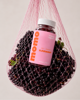 A bottle of momo elderberry supplement with a pink label, nestled inside a pink mesh bag filled with fresh dark elderberries, set against a solid light beige background.