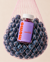A bottle of LION'S MARE blueberry flavor gummies and cranberry sauce, featuring a purple label with orange 'momo' branding, nestled inside a pink mesh bag filled with blueberries against a solid beige background.