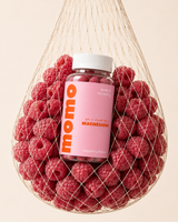 A bottle of momo brand magnesium supplement with a pink label and white cap, surrounded by fresh red raspberries inside a beige mesh bag on a light beige background.