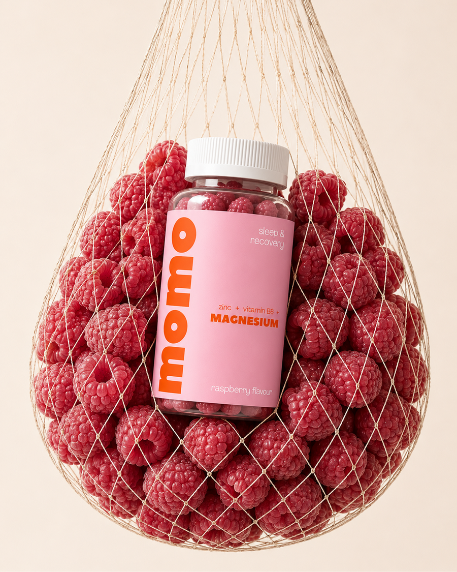 A bottle of momo brand magnesium supplement with a pink label and white cap, surrounded by fresh red raspberries inside a beige mesh bag on a light beige background.