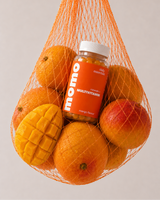 A bottle of momo multivitamin gummies nestled inside an orange mesh bag filled with mangoes and oranges, photographed against a clean light gray background.