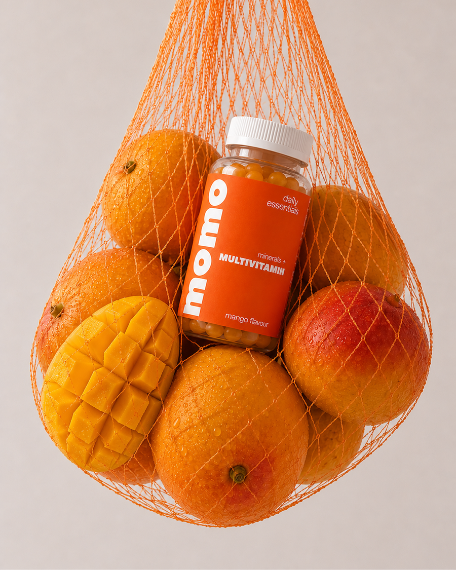 A bottle of momo multivitamin gummies nestled inside an orange mesh bag filled with mangoes and oranges, photographed against a clean light gray background.