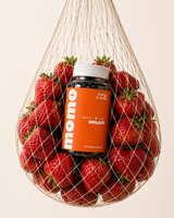 A bottle of MOMO Shilajit vitamin and mineral supplement, surrounded by fresh strawberries inside a hanging mesh bag, on a solid light beige background.