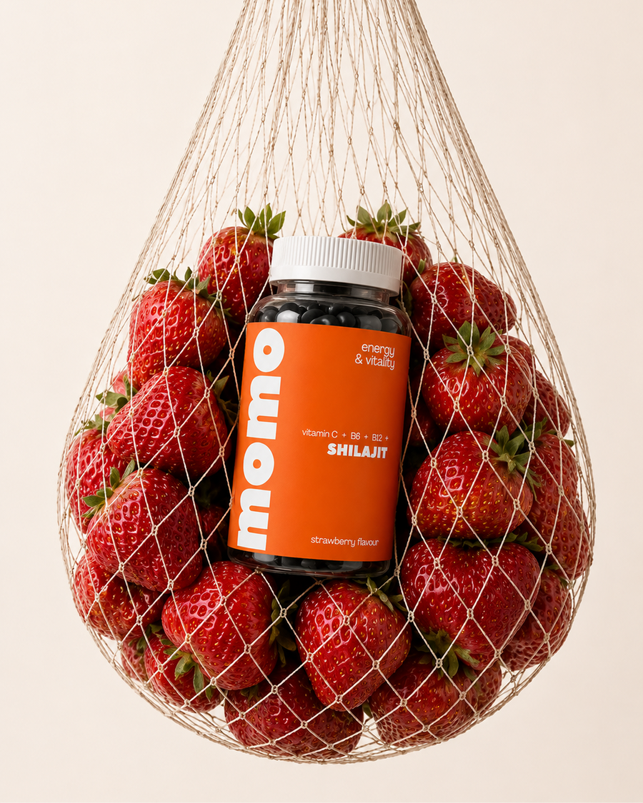 A bottle of MOMO Shilajit vitamin and mineral supplement, surrounded by fresh strawberries inside a hanging mesh bag, on a solid light beige background.